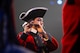 An Army Soldier dressed in Revolutionary War-era red uniform with black tri-cornered hat is playing a fife.