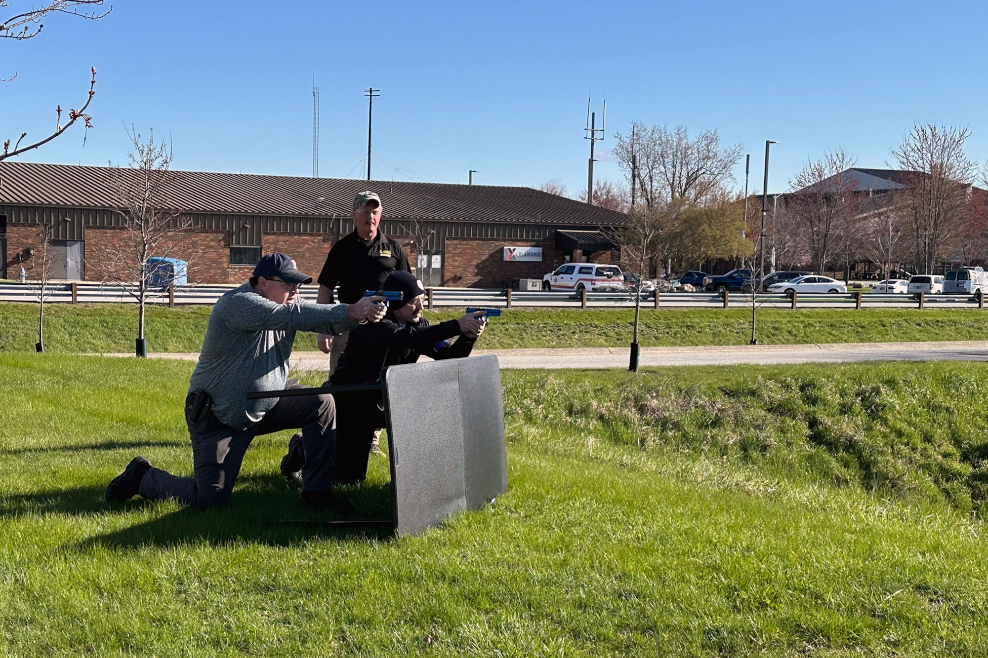 Two law enforcement personnel stand behind barricade with training weapons aimed, with an observer in background.