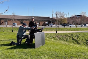 Two law enforcement personnel stand behind barricade with training weapons aimed, with an observer in background.