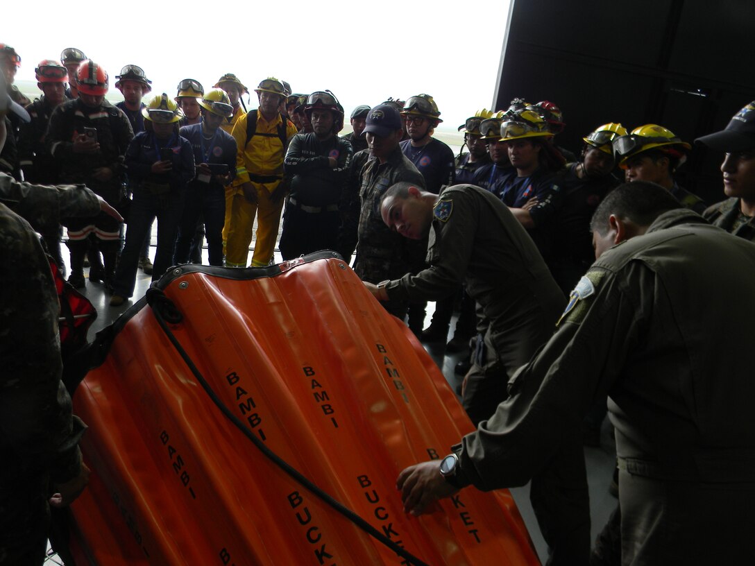 A group of firefighters and solider look at a water-carrying basket designed to be suspended from helicopters during training.