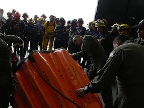 A group of firefighters and solider look at a water-carrying basket designed to be suspended from helicopters during training.
