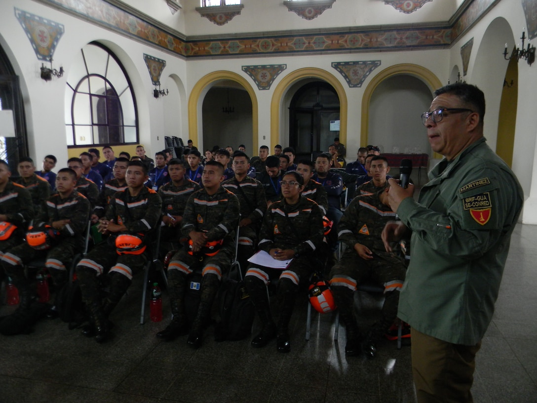 A man in a green uniform stands in front of a large group of sitting firefighters and speaks.