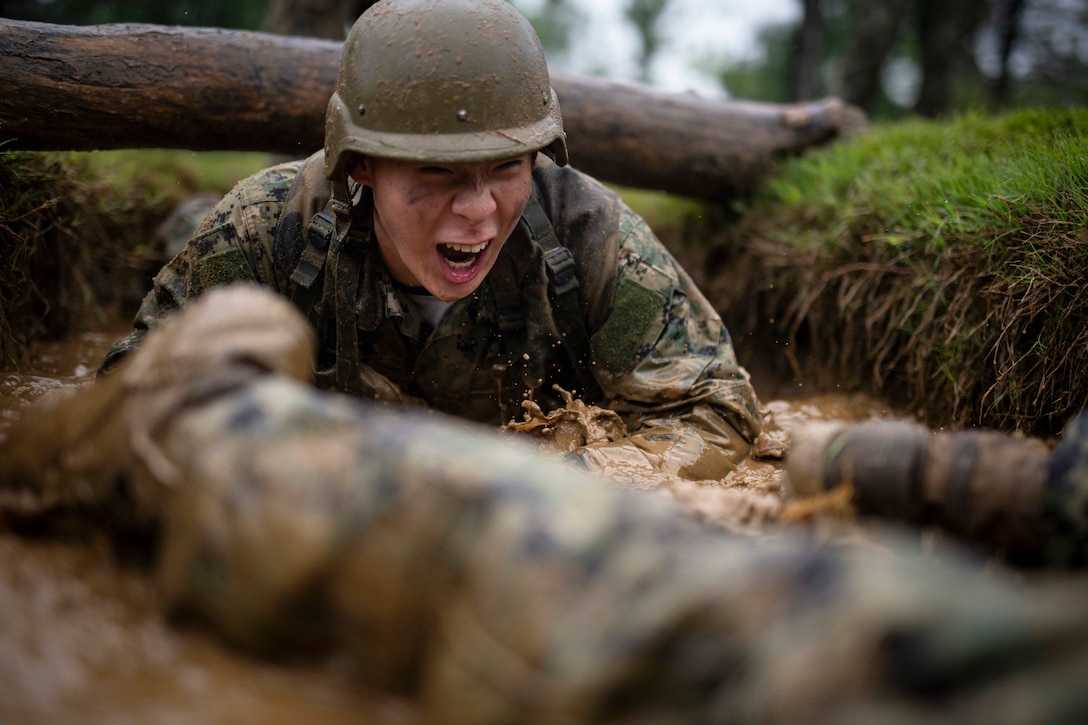 A U.S. Naval Academy midshipman climbs through the mud under a log in a wooded area on a gloomy day.