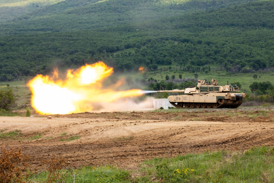 A tank fires a round creating an orange explosion on a dirt field with greenery in the background.