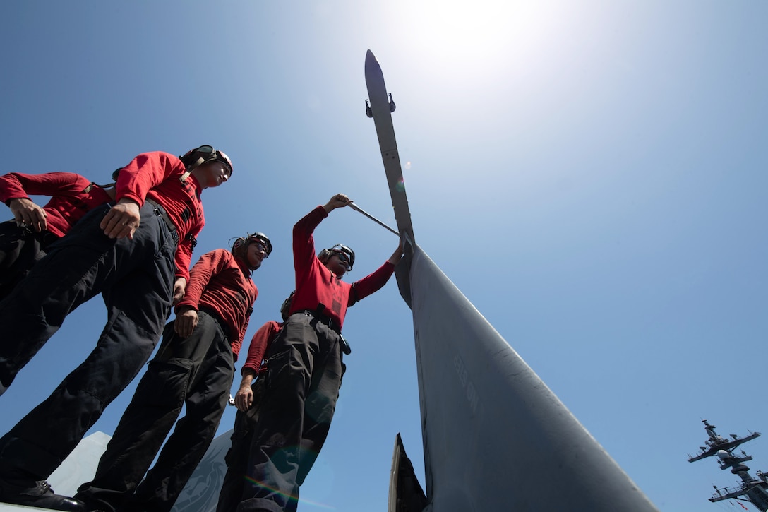 Five sailors in red gear work on a ship against a blue sky as seen from below.