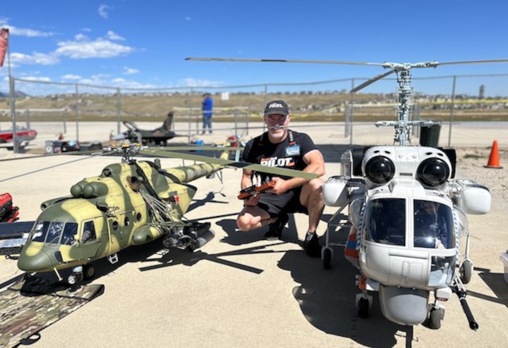 A gentleman kneeling in between two scale, radio-controlled helicopters.