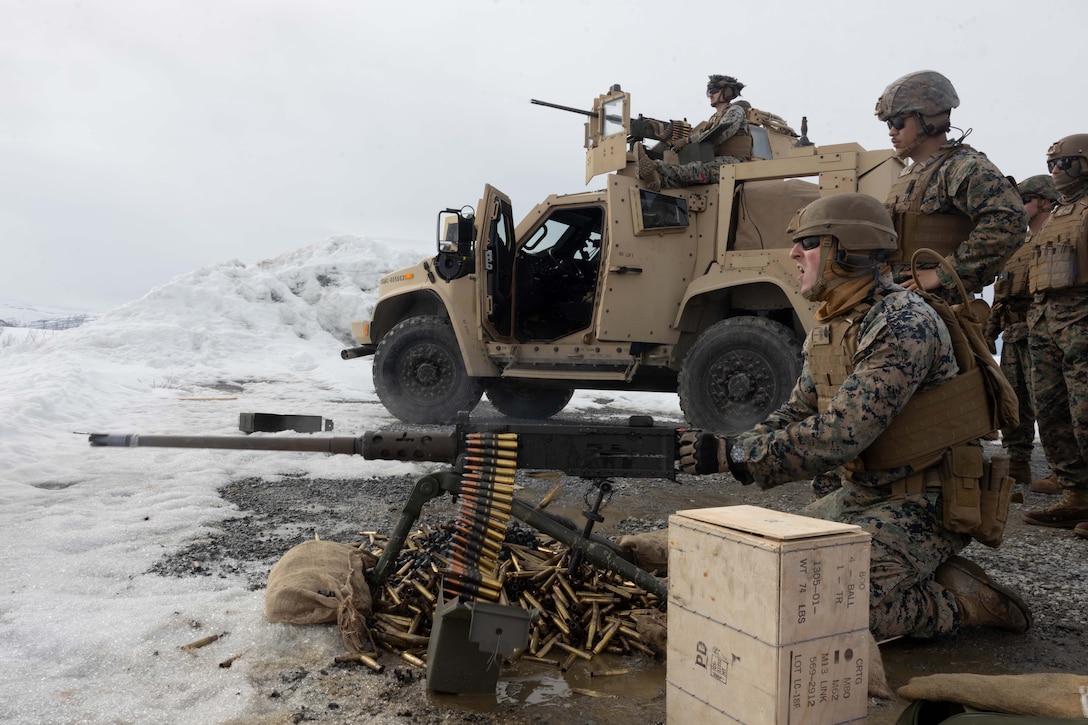 A Marine fires a machine gun while other Marines watch on and near a vehicle in the snow.