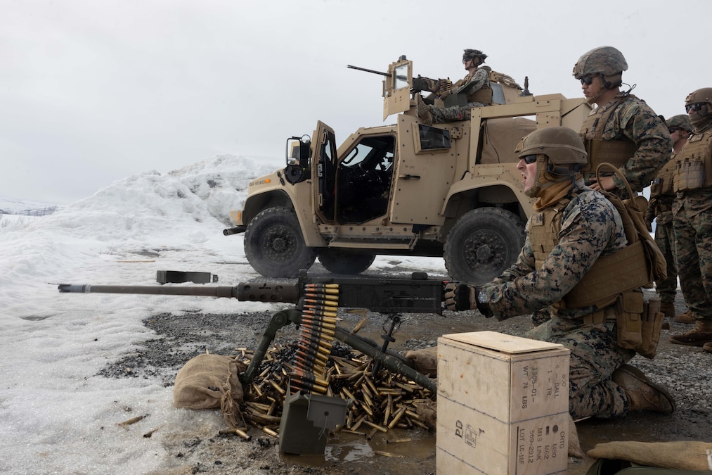 A Marine fires a machine gun while other Marines watch on and near a vehicle in the snow.