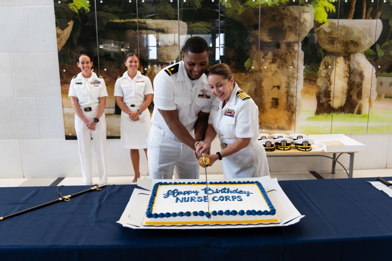 Two military personnel in dress uniforms use a saber to cut a cake that reads "Happy Birthday Nurse Corps."