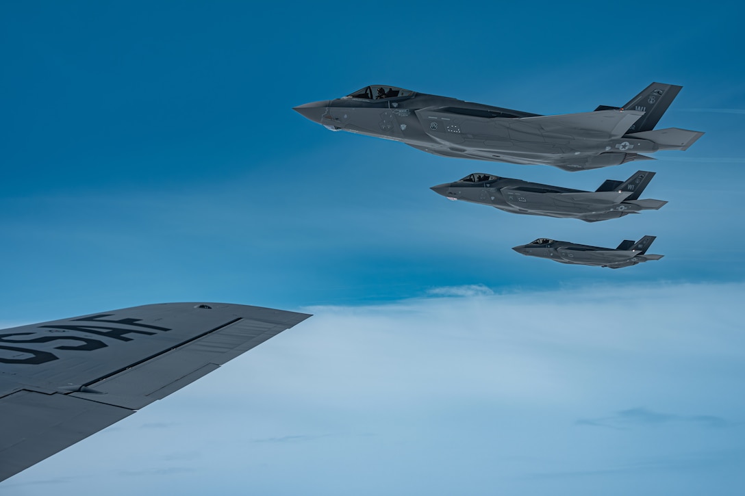 Three aircraft fly in formation next to another aircraft wing above clouds in a blue sky.