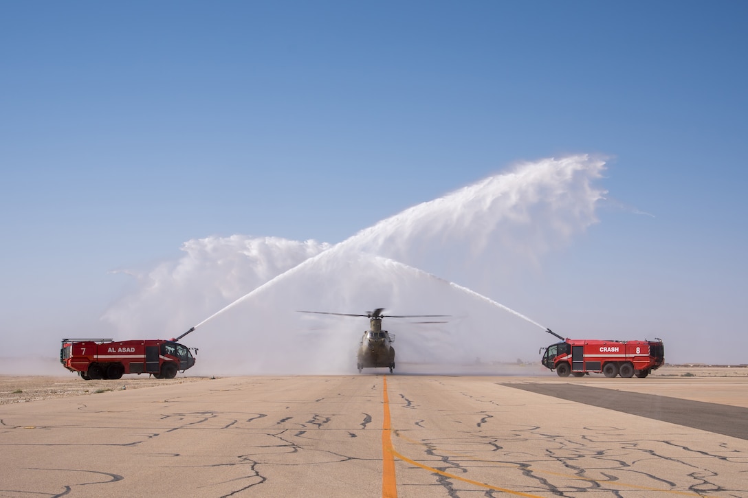 Two red vehicles spray water over a helicopter on a runway under blue skies.