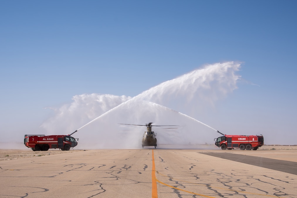 Two red vehicles spray water over a helicopter on a runway under blue skies.