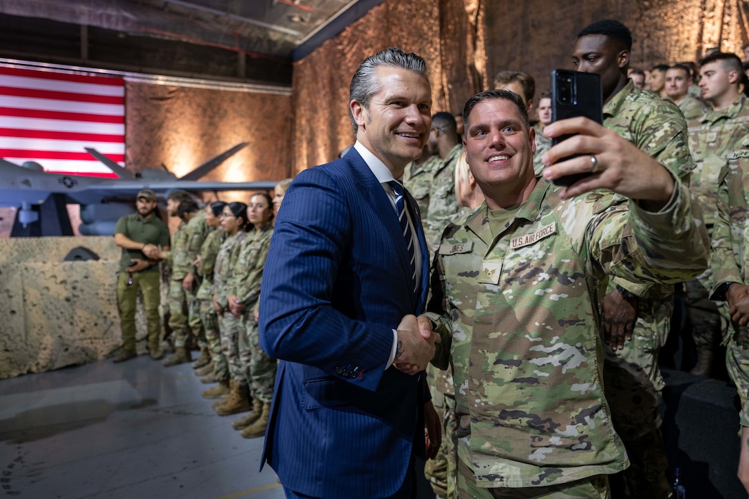 A man in a suit takes a selfie with an airman in uniform in the front row of a group of airmen standing on bleachers in an auditorium.
