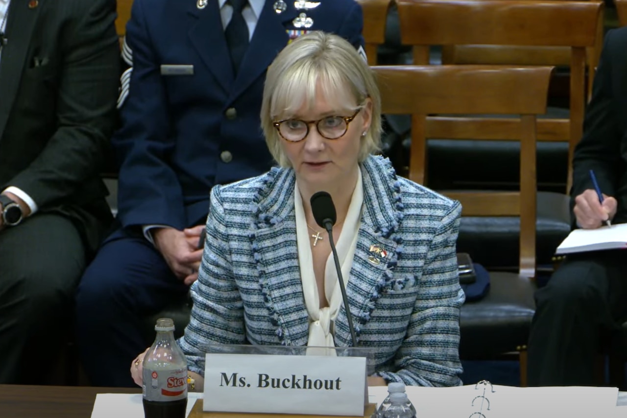 A woman in business attire sits at a table and speaks into a microphone. A man in a military dress uniform and two in business attire sit behind her.