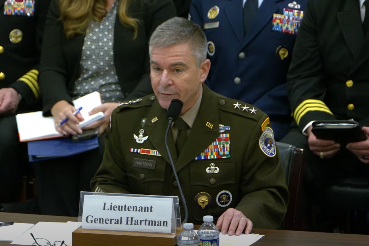 A man in a military dress uniform sits at a table and speaks into a microphone. Several men in military dress uniforms and a woman in business attire sit behind him.