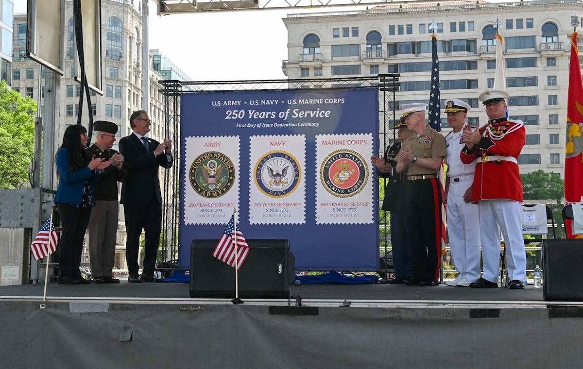 Seven individuals in military dress uniforms and business attire stand and clap in front of a display that shows three postage stamps.