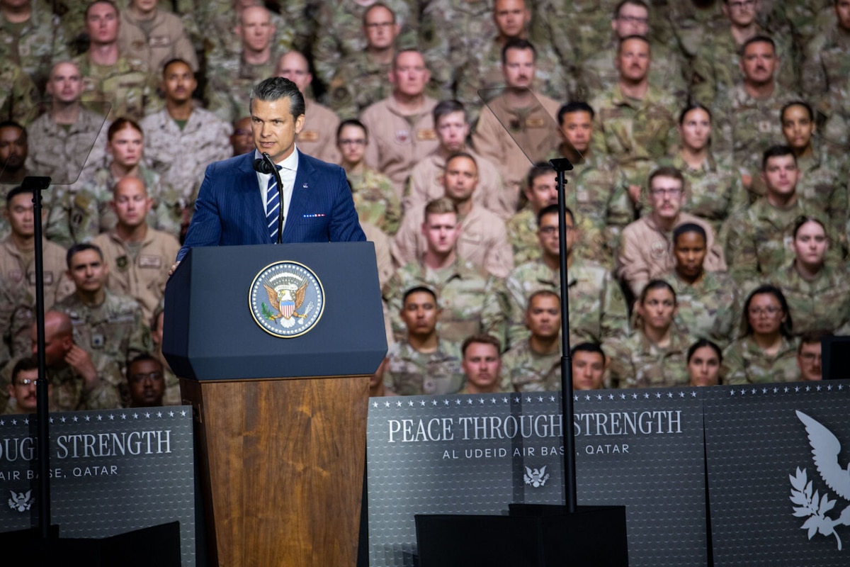 A man in a suit stands behind a lectern, and dozens of uniformed military personnel stand behind him.