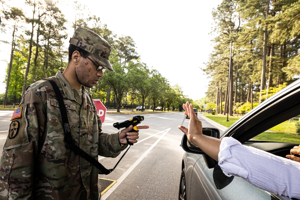A man wearing a camouflage military uniform holds and scans an ID card for a person entering a military installation. Across from him is a person in a car with an arm outstretched and holding a card. A tree-lined road is in the background.