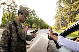 A man wearing a camouflage military uniform holds and scans an ID card for a person entering a military installation. Across from him is a person in a car with an arm outstretched and holding a card. A tree-lined road is in the background.