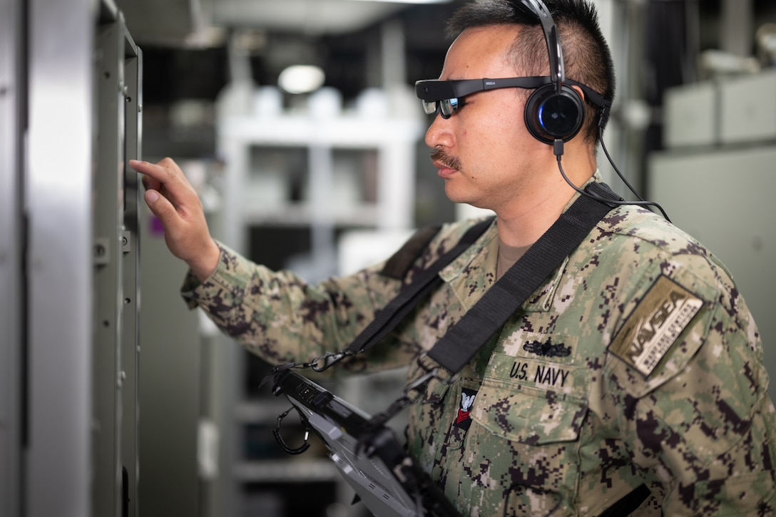 A sailor wearing a headset and augmented reality glasses touches a device on a ship wall.