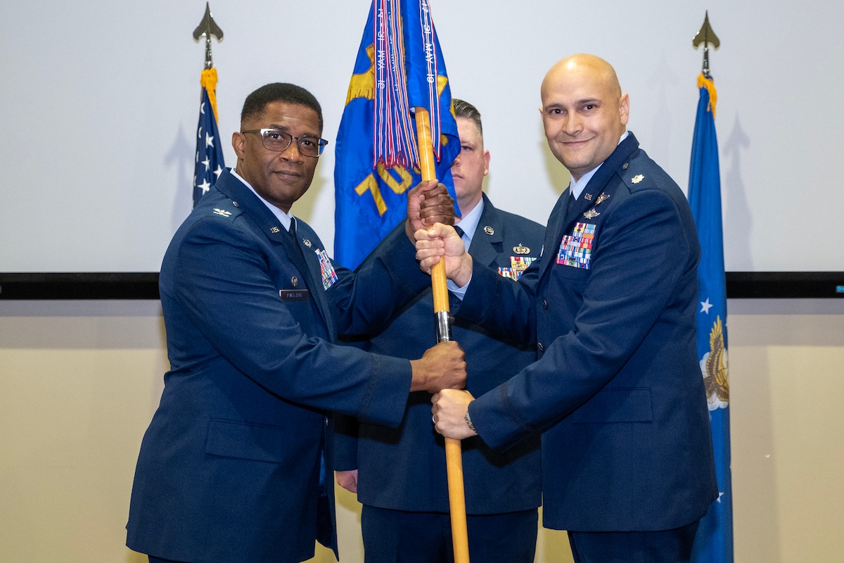 photo of three uniformed US Air Force Airmen standing on a stage, two holding a unit flag while one stands at attention behind them.