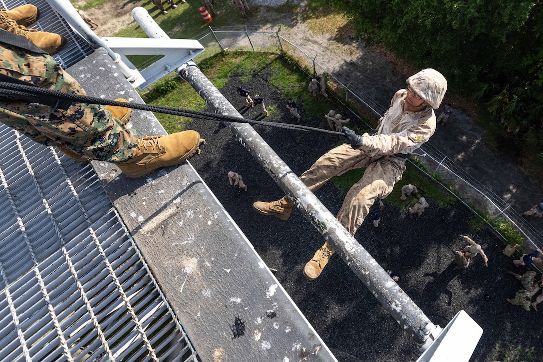 A Marine Corps recruit rappels down a tower with safety on gear on as fellow recruits gather on the ground as seen from above.