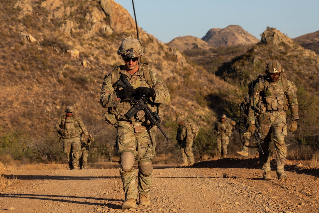Soldiers in combat gear and carrying rifles walk along a dirt road with mountains in the background.