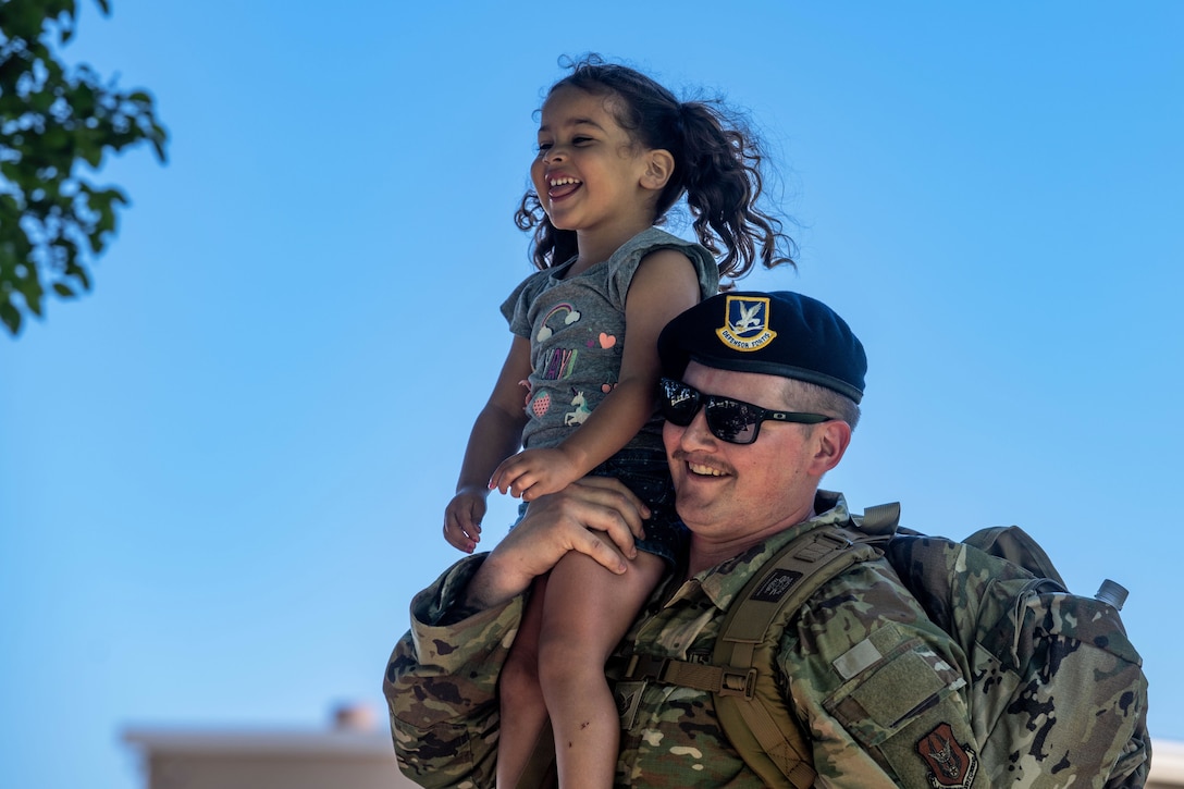 A close-up of a smiling airman carrying a child on their shoulders against a blue sky.