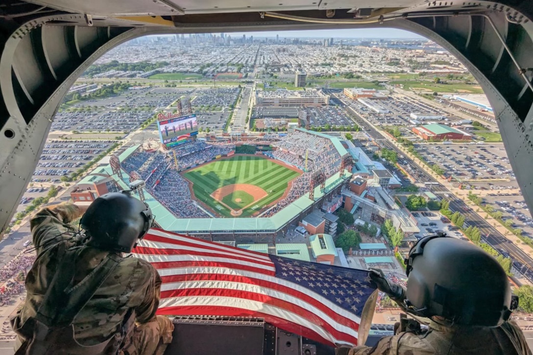Two soldiers in tactical gear sit in the open doorway of an airborne helicopter while displaying an American flag over a baseball stadium.