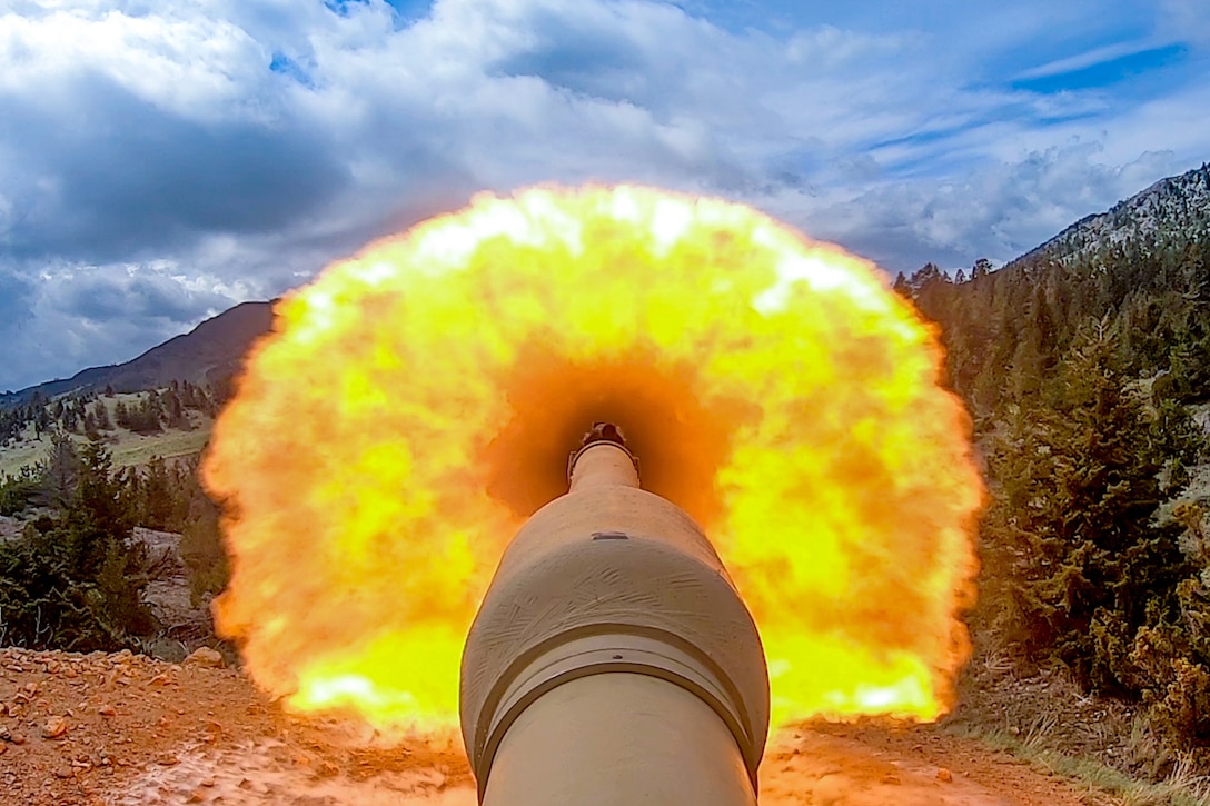 A close-up of a tank's fume extractor and barrel as it fires creating a large orange fireball toward mountains covered with trees under a cloudy, blue sky.