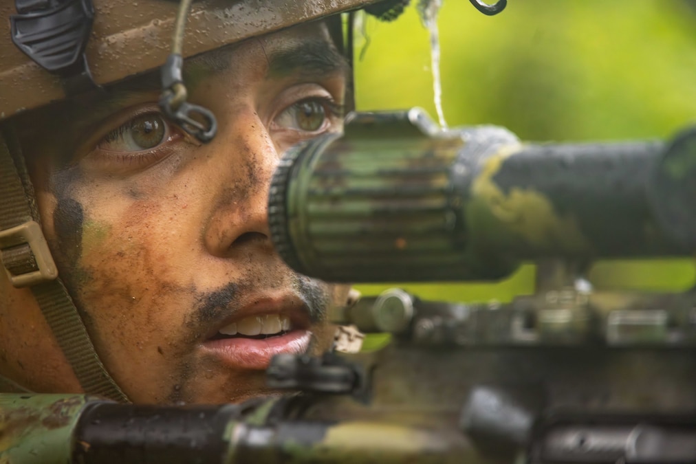 A Marine holding a rifle with a sight device on top looks into the distance.