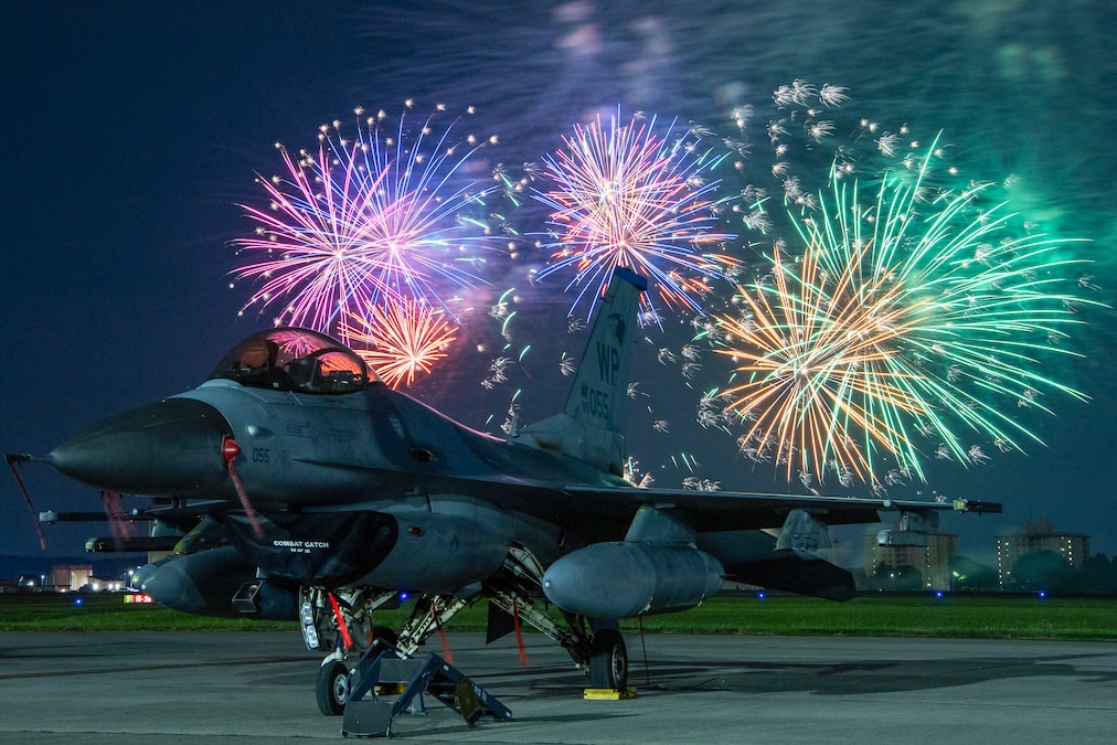 A fighter jet is parked on a runway as fireworks light the sky behind it.