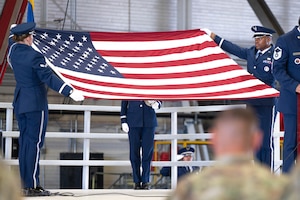 Four Airmen stand on stage during a Police Week ceremony at Scott Air Force Base. One Airman stands at a podium while three honor guardsmen perform Taps; two of them hold a fully extended American flag.