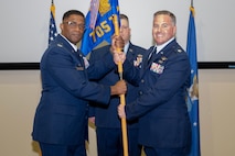 photo of three uniformed US Air Force Airmen standing on a stage, two holding a unit flag while one stands at attention behind them.