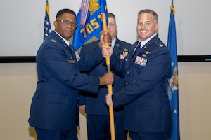 photo of three uniformed US Air Force Airmen standing on a stage, two holding a unit flag while one stands at attention behind them.