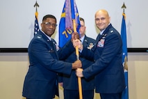 photo of three uniformed US Air Force Airmen standing on a stage, two holding a unit flag while one stands at attention behind them.
