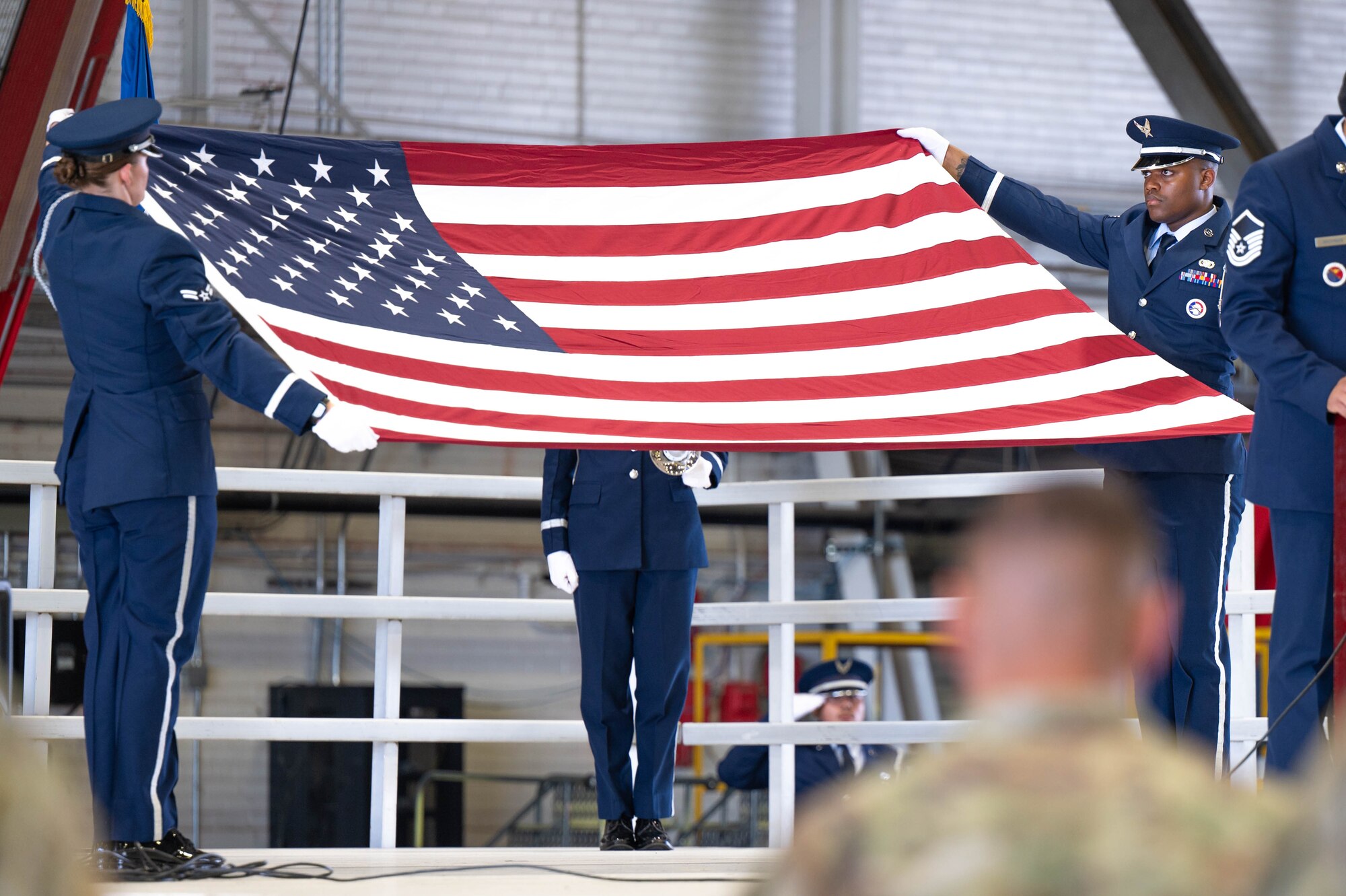 Four Airmen stand on stage during a Police Week ceremony at Scott Air Force Base. One Airman stands at a podium while three honor guardsmen perform Taps; two of them hold a fully extended American flag.