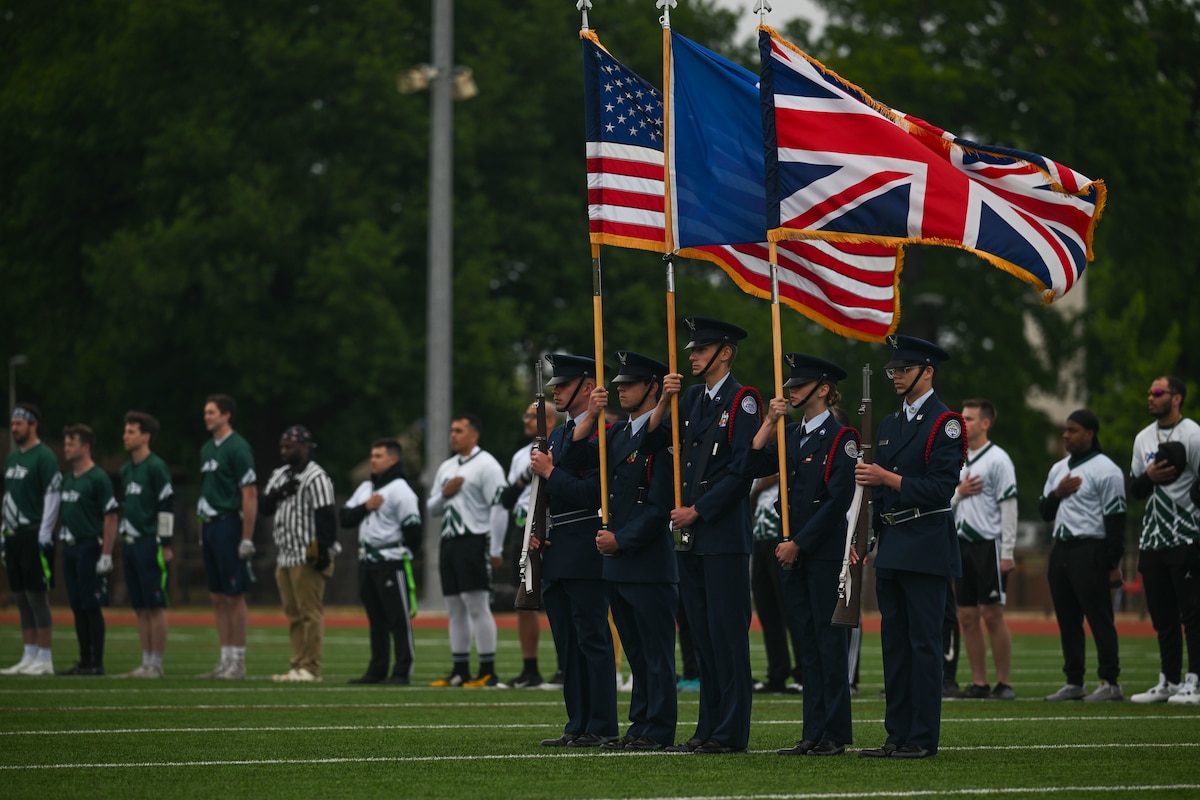 Five people wearing formal military uniforms hold the American, the U.S. Air Force, and the United Kingdoms flags on a football field with a long row of players holding their hands over their hearts in the background