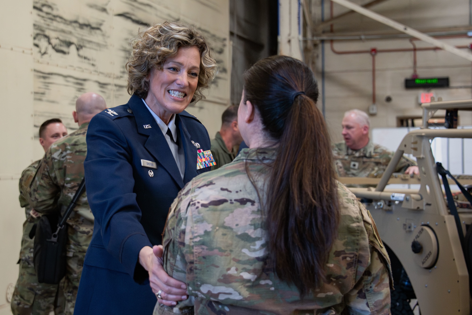 A woman greets another by touching a hand to her shoulder and smiling.