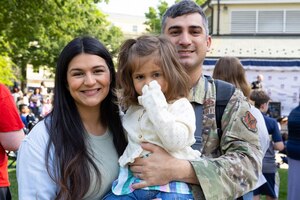 A man in a military camouflage unifrom and a woman in casual attire hold a child. There are several people in casual attire in the background.