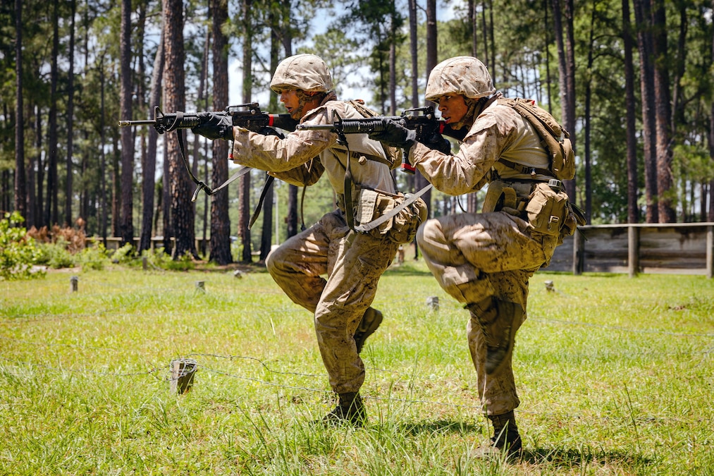 Two Marines aim weapons in a field while both standing with one leg lifted and bent at the knee.