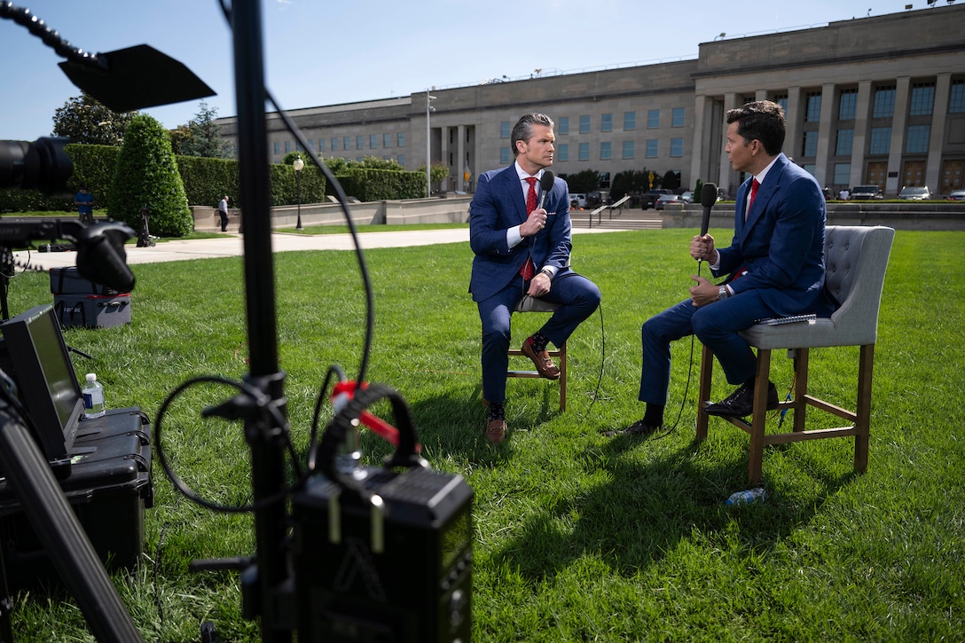 The defense secretary and another civilian hold microphones and sit in chairs facing each other on the lawn outside the Pentagon.