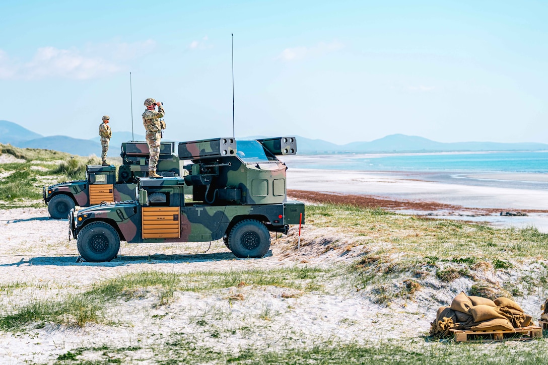 Two soldiers stand on vehicles parked in the sand near the beach while looking through binoculars with mountains in the distance.