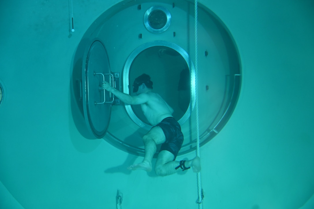 A close-up of a sailor wearing goggles swims into a partially visible submarine training device.