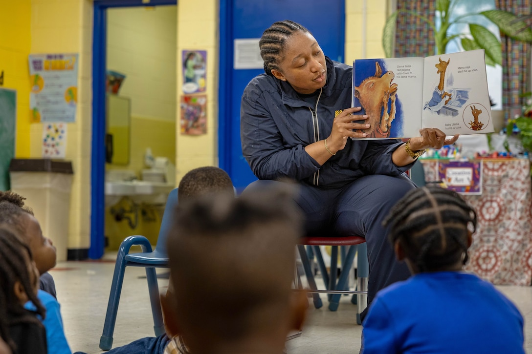 A sailor reads a book to children in a yellow and blue classroom.