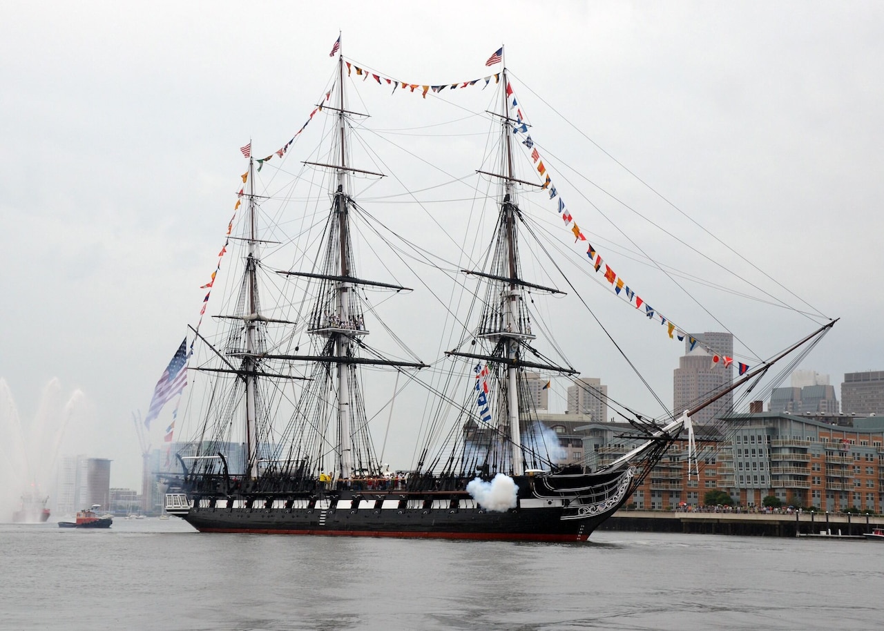 A sailing ship in a harbor fires its cannons.