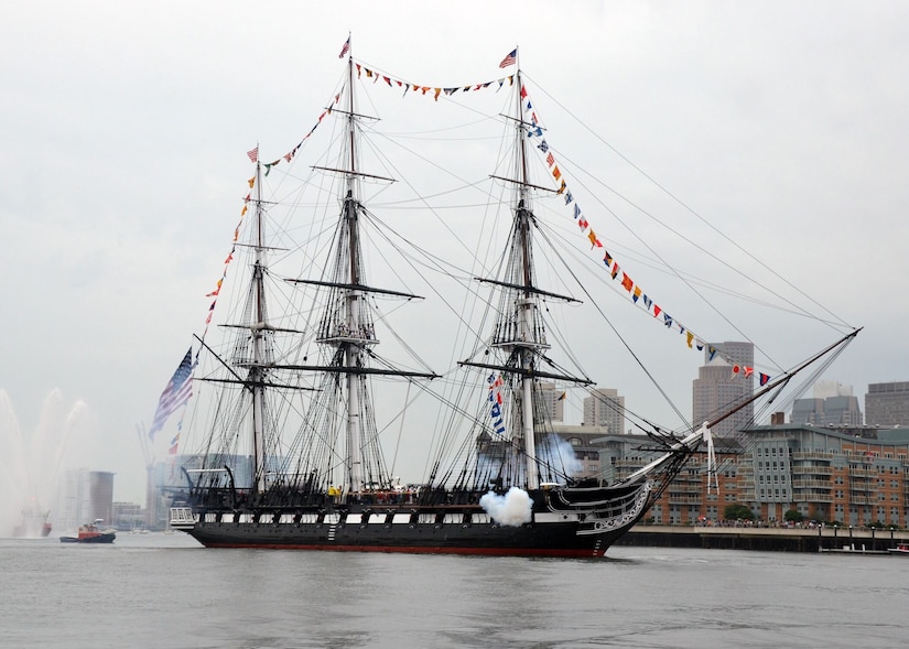 A sailing ship in a harbor fires its cannons.