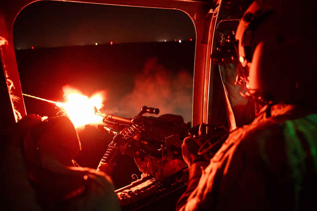 A soldier in an aircraft fires a machine gun into the night sky.