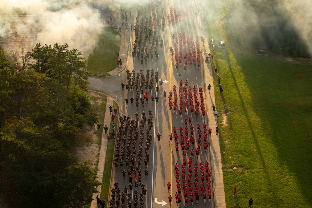An elevated view of a large group of soldiers running on a paved street as onlookers watch from each side.