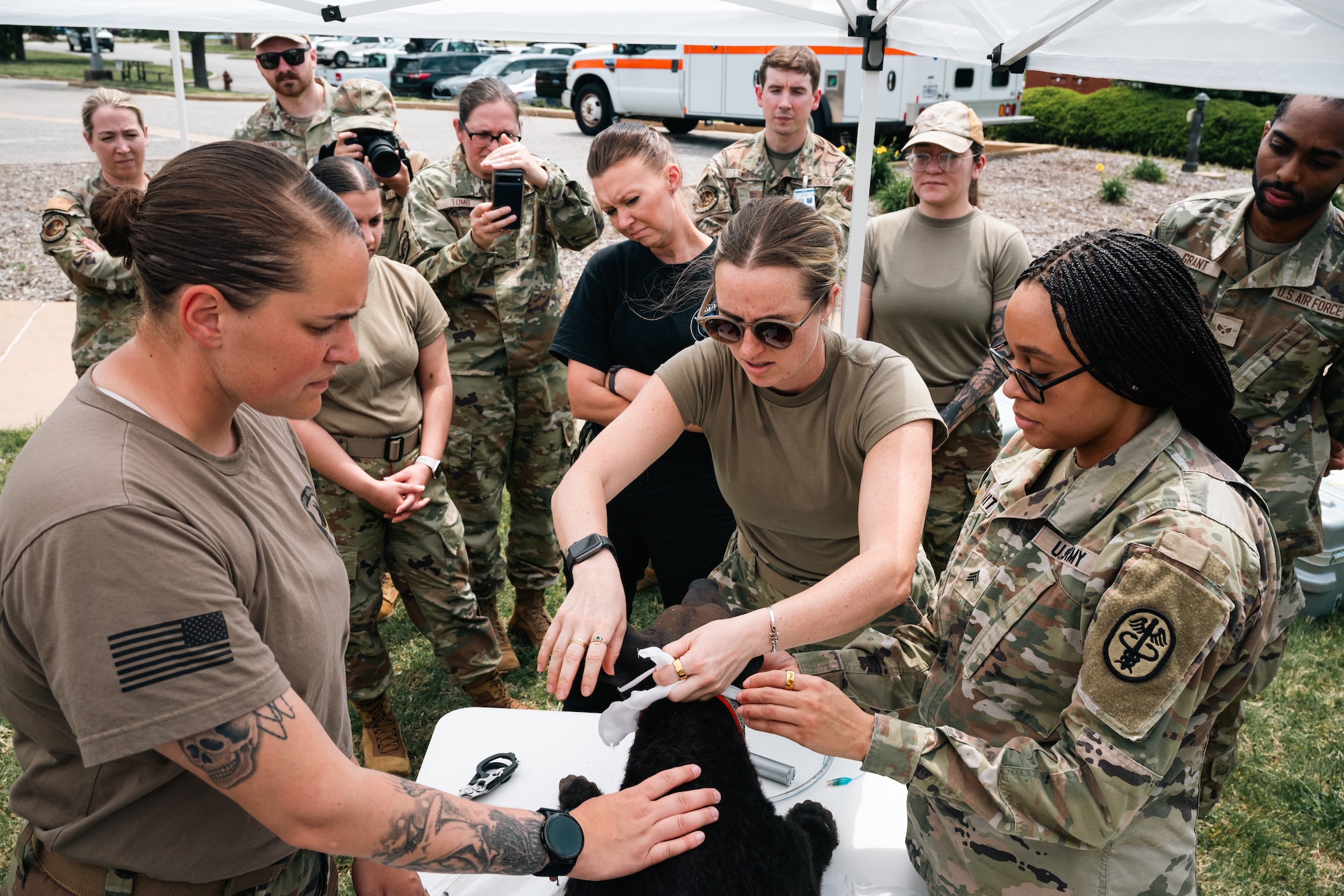 A group of Airmen and Soldiers gather around a table with a medical training dog dummy on a white table outside